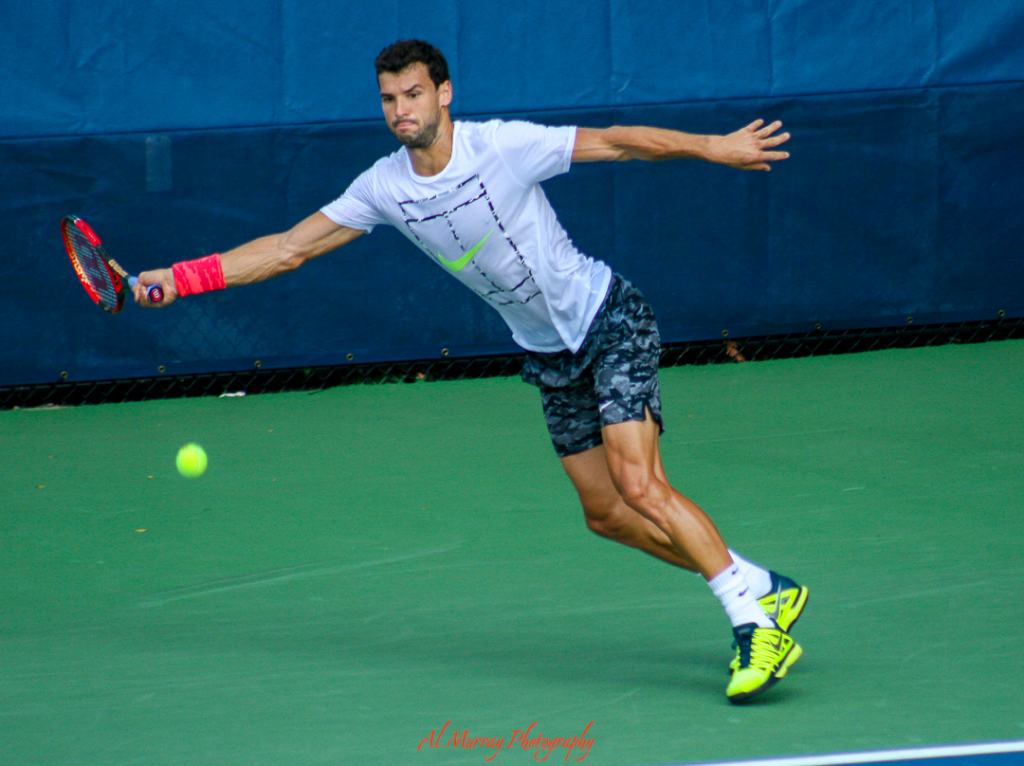 US Open travel guide for older tennis fans Male tennis player stretches to hit a forehand shot on a green hard court with a blue tarp backdrop, typical of the practice courts at the US Open. His focused posture and athletic gear highlight the intensity and preparation that happens behind the scenes.