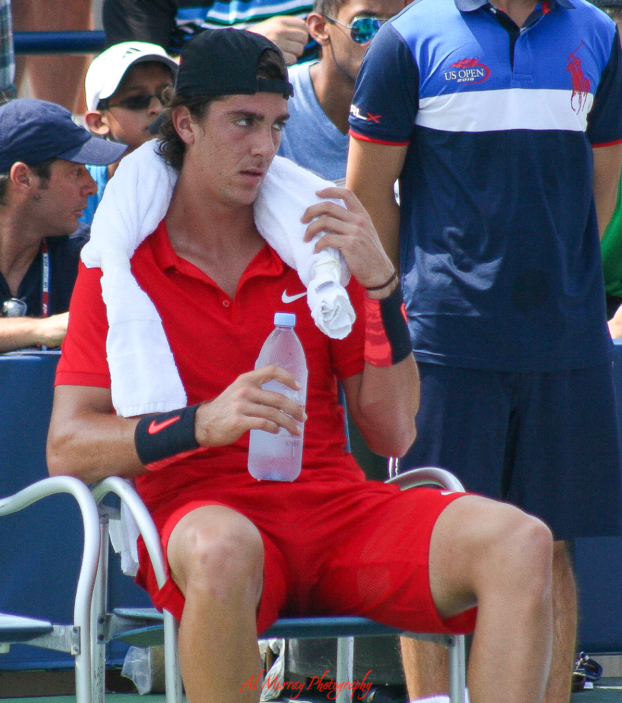 US Open travel guide for older tennis fans Male tennis player in a red Nike outfit sits courtside at the US Open, cooling off with a towel around his neck and holding a bottle of water. His focused expression and the official US Open staff shirt in the background capture the intensity and heat of match day.