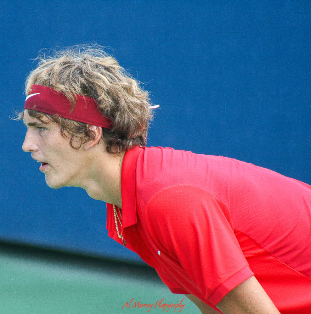 US Open travel guide for older tennis fans Young male tennis player in a red Nike outfit and matching headband intensely focuses before receiving serve on a hard court at the US Open. His ready stance reflects one of the essential US Open tennis tips: stay alert and anticipate every point.