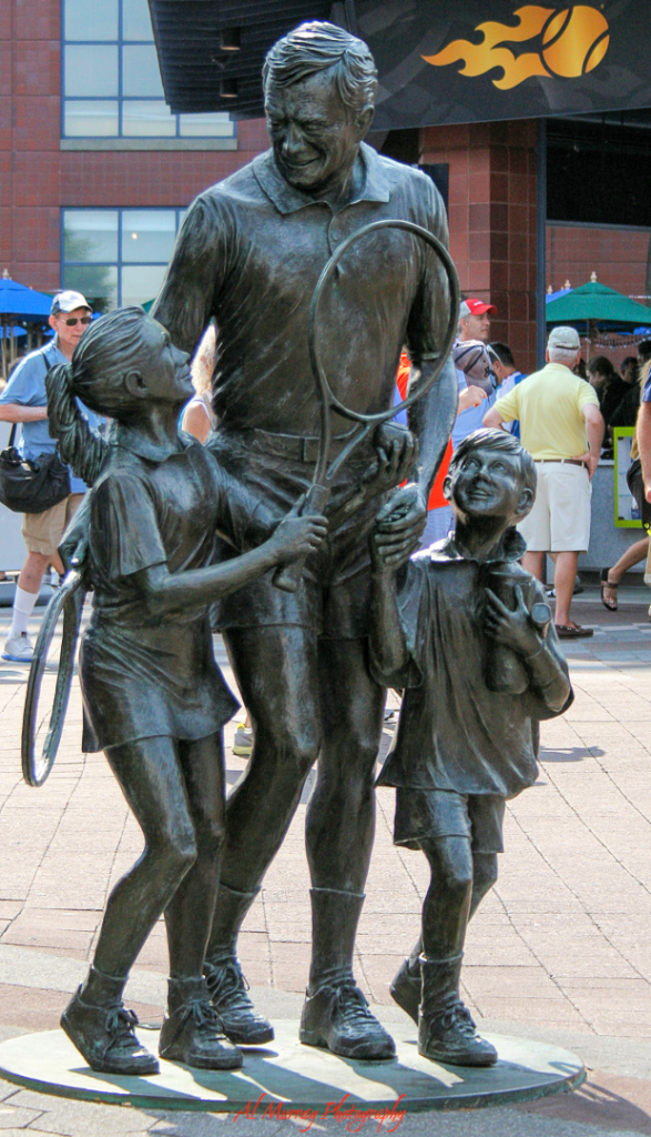 US Open travel guide for older tennis fans Bronze statue of a smiling man holding tennis rackets while engaging with two young children, located outside the Billie Jean King National Tennis Centre. This welcoming sculpture symbolizes the family-friendly spirit and tennis legacy celebrated at the venue.