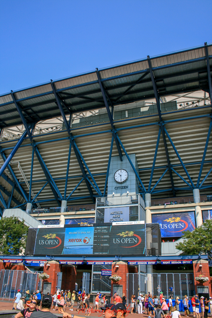 US Open travel guide for older tennis fans Wide-angle view of Arthur Ashe Stadium at the US Open, featuring the iconic Citizen clock, digital screens displaying event info, and banners with the tournament’s flaming tennis ball logo. Fans gather at the entrance beneath the “Night Session Entry” signs, capturing the excitement of visiting Arthur Ashe Stadium at the US Open.