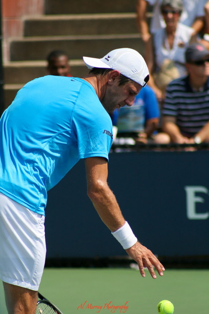 Male tennis player in a blue shirt and white cap tosses a ball to serve on a hard court, with spectators watching from tiered seating in the background. Captures the atmosphere of daytime matches, offering a tip for avoiding crowds—opt for outer courts or earlier rounds.