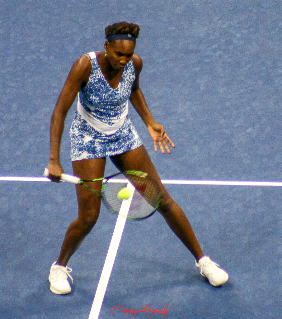 US Open travel guide for older tennis fans A female tennis player in a blue and white patterned outfit returns a low shot on a hard court during a US Open match. The action shot captures the intensity and athleticism fans can expect when attending the US Open.