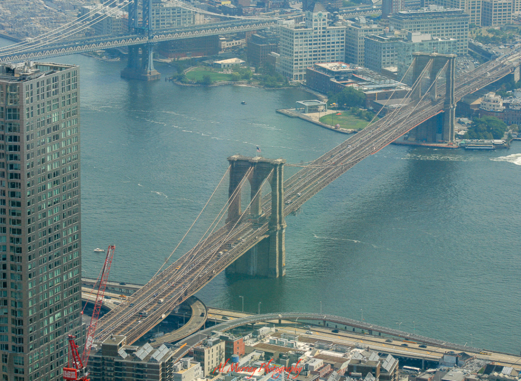 Aerial view of the Brooklyn Bridge stretching across the East River with the Manhattan Bridge in the background, surrounded by city buildings and calm waters. A breathtaking sight that reminds travelers some iconic experiences—like walking this historic span—are truly worth the effort.