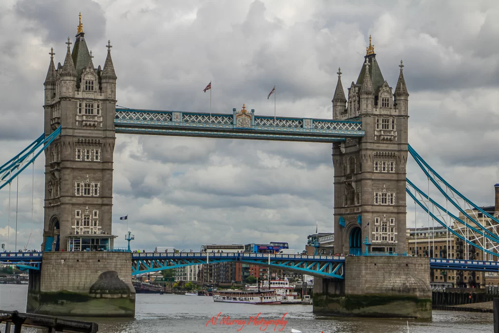 Tower Bridge in London with its twin stone towers and iconic blue suspension elements spanning the River Thames under a cloudy sky. A must-see historic landmark featured in a Wimbledon guide for mature travellers seeking classic London experiences.