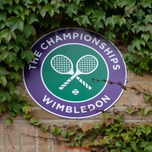The Championships Wimbledon logo featuring two crossed tennis rackets on a green and purple circular sign mounted on a brick wall covered in ivy. A timeless emblem included in a Wimbledon guide for mature travellers seeking tradition and elegance.
