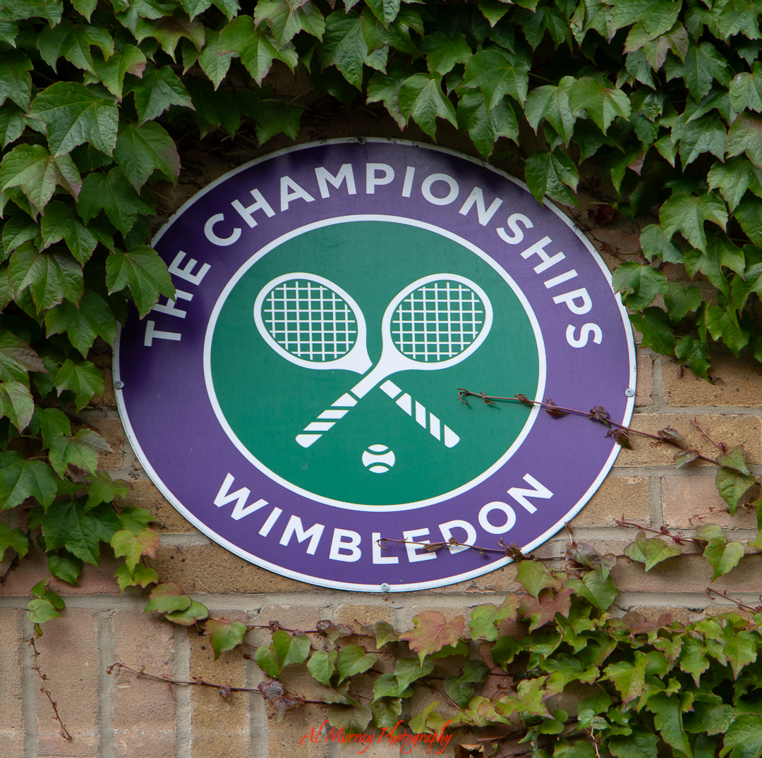 The Championships Wimbledon logo featuring two crossed tennis rackets on a green and purple circular sign mounted on a brick wall covered in ivy. A timeless emblem included in a Wimbledon guide for mature travellers seeking tradition and elegance.