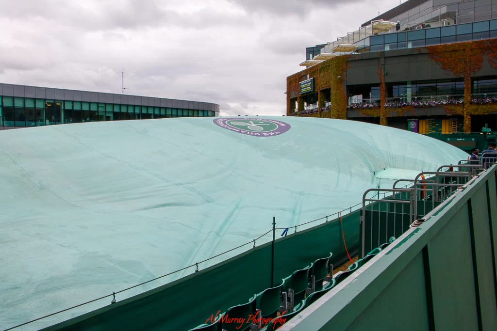 Tennis court at Wimbledon covered with a large green tarp bearing the tournament’s logo, with empty spectator seats and overcast skies above. This image captures the familiar pause caused by rain at Wimbledon.
