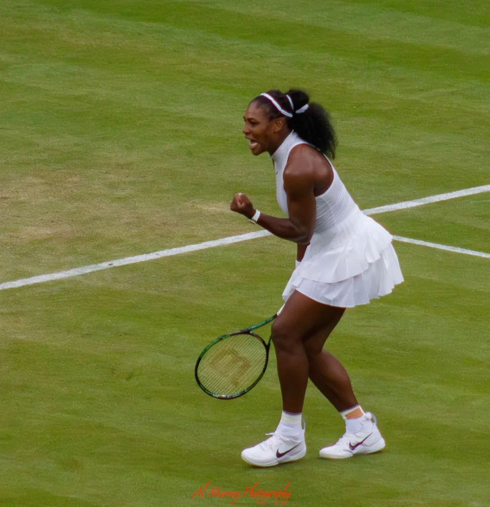Female tennis player in a white outfit clenching her fist and celebrating a point on a grass court, showing fierce determination during a match. A powerful moment from a grand slam tournament, capturing the intensity of elite competition.
