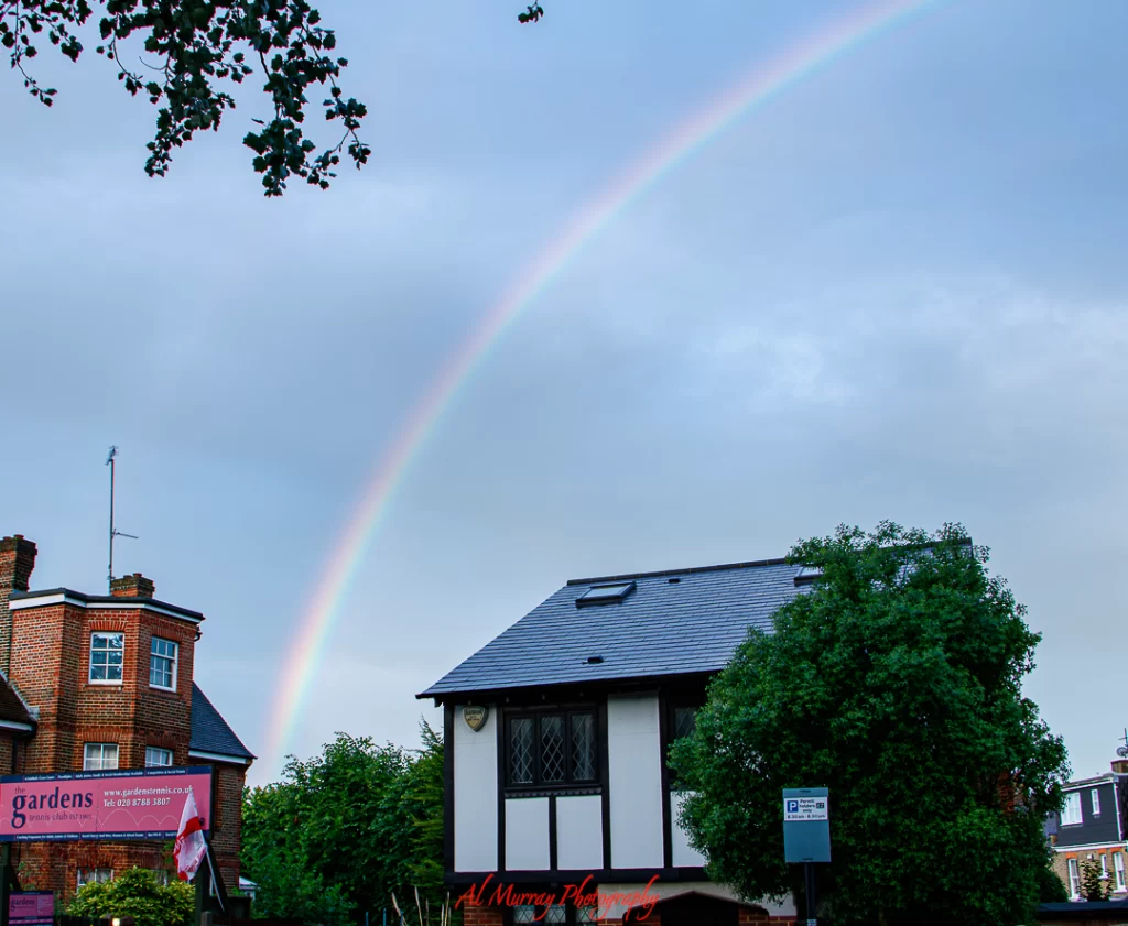 A soft rainbow arcs across a cloudy sky above charming residential buildings in Wimbledon, including a sign for The Gardens Tennis Club. A serene moment capturing the beauty of a rainbow over Wimbledon.