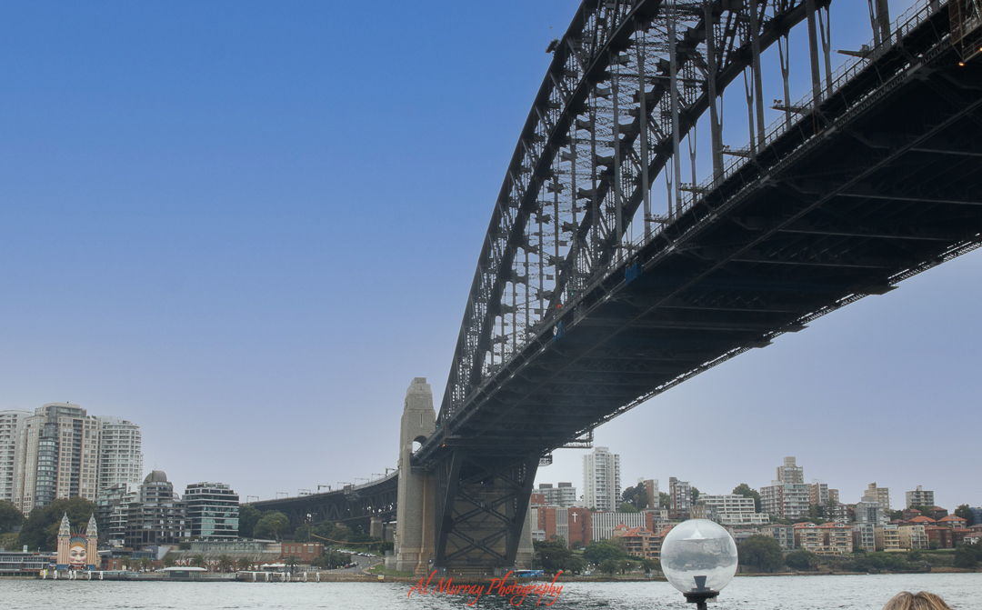 Wide-angle view of the Sydney Harbour Bridge with city buildings in the background and waterfront in the foreground, capturing the iconic Australian setting that enhances the Australian Open experience for older tennis fans visiting Sydney. The clear sky and urban landscape add to the scenic charm of the destination.
