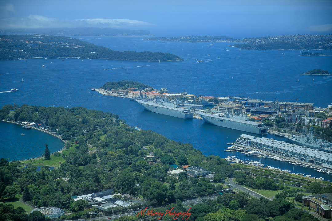 Aerial view of Sydney Harbour featuring naval ships docked near Garden Island, surrounded by lush greenery, marinas, and city buildings under clear blue skies. A scenic destination ideal for mature travellers seeking a blend of coastal beauty, maritime history, and relaxed exploration.