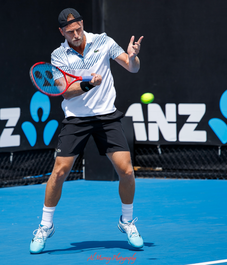 Senior male tennis player mid-air during a forehand swing at a professional tournament, wearing a striped white polo, black shorts, and light blue tennis shoes on a blue hard court. The background features ANZ branding, suggesting a formal event setting suitable for competitive senior athletes.