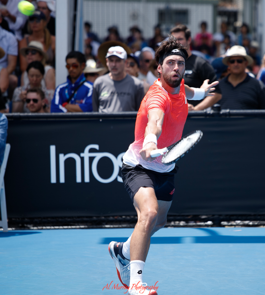 Professional male tennis player in mid-swing during a daytime match on a blue hard court, with a focused crowd watching closely behind him. Captures the live excitement fans can expect when planning a tennis tournament trip to high-energy venues.