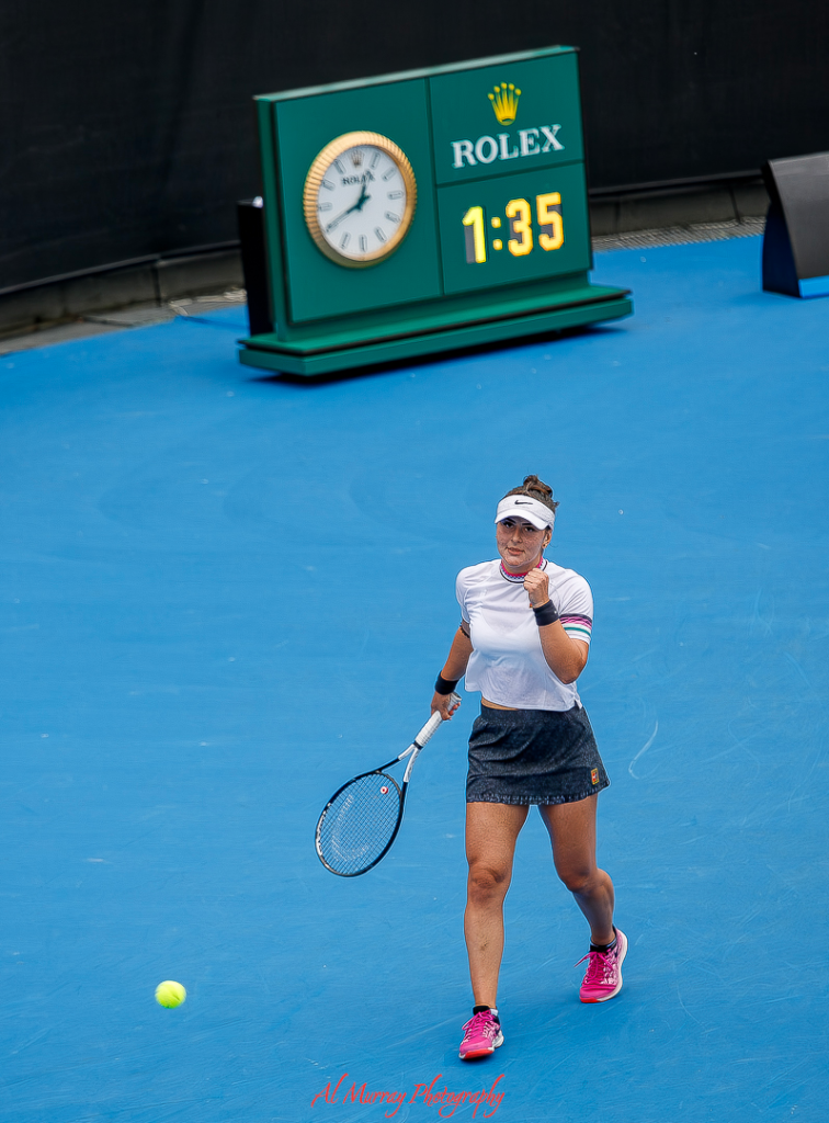Female tennis player pumps her fist in celebration on a bright blue court at the Australian Open, with a Rolex clock and scoreboard in the background reading 1:35. Her focused expression and dynamic posture capture the energy of one of the best Australian Open experiences for fans and players alike.