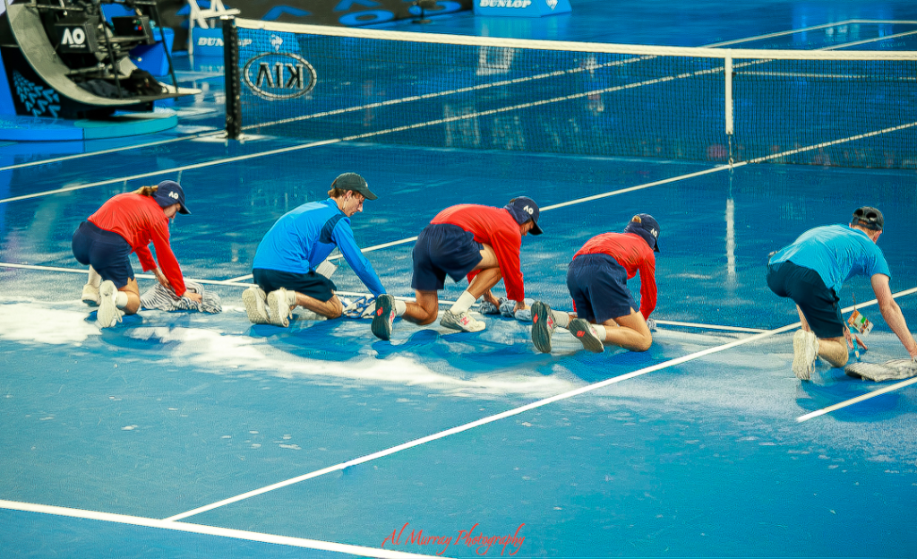 Five ball kids in red and blue uniforms mop up rainwater on a bright blue tennis court during a break in play at the Australian Open. Their synchronized effort shows how quickly the tournament responds to weather delays, offering a behind-the-scenes look relevant to Australian Open tips.