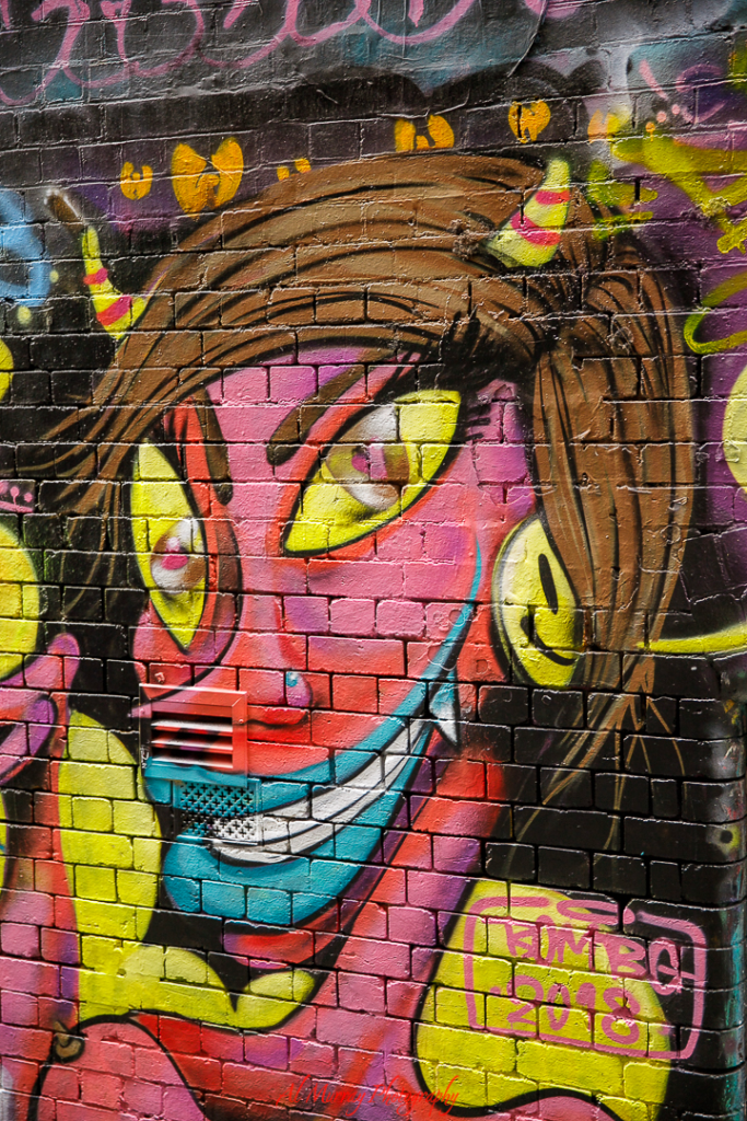 Vivid street art mural of a grinning cartoon-like character with bright pink skin, yellow eyes, and colorful horns, painted on a brick wall in an urban alley. A playful visual moment seniors might encounter while exploring off-the-beaten-path spots on a tennis travel adventure.