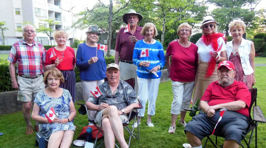 Group of seniors gathered outdoors on a green lawn, smiling and holding Canadian flags, some seated in lawn chairs and others standing, dressed casually in red and white. While the image shows a festive Canada Day celebration, it evokes the sense of community that solo seniors in Canada can seek out during holidays like Valentine’s Day.

