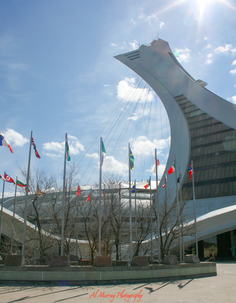 The Montreal Tower at Olympic Park rises dramatically into a bright sky, with international flags waving in the foreground and bare winter trees surrounding the plaza. This iconic landmark adds a unique and adventurous stop to a Montreal Valentine's Day itinerary for couples.
