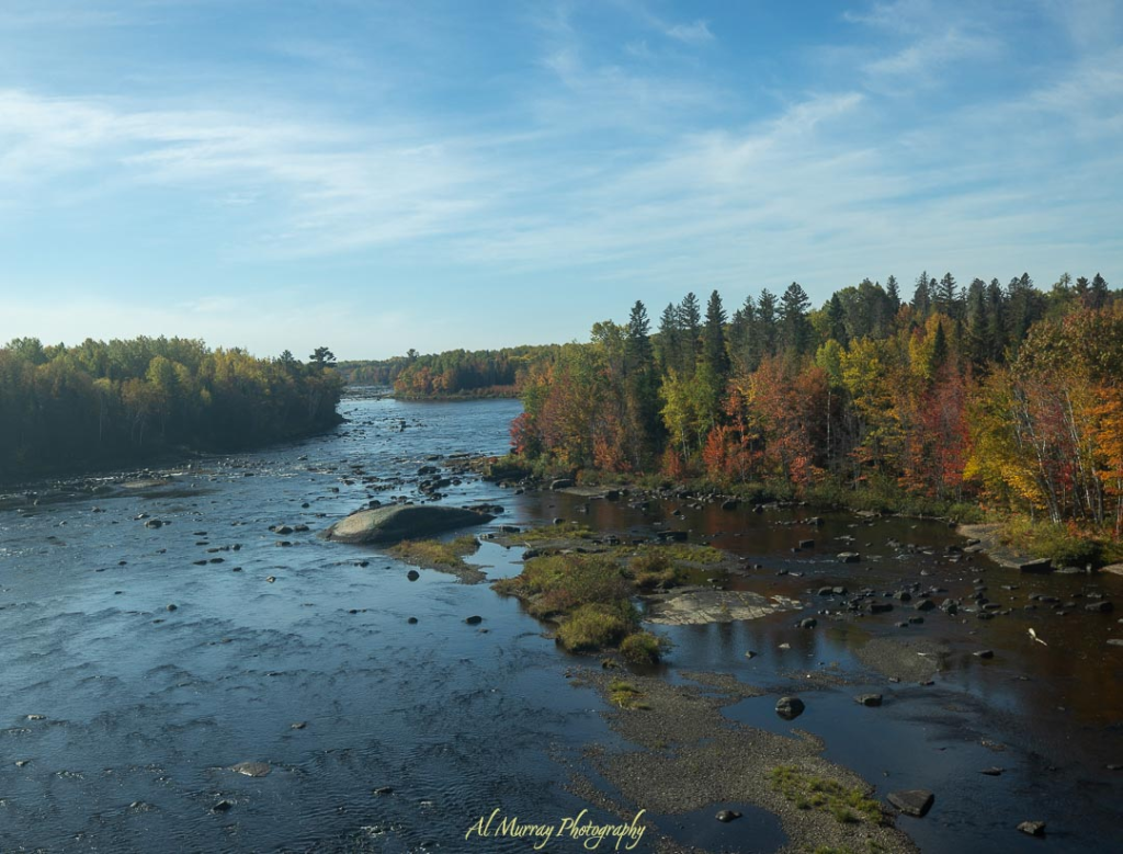 Scenic river bordered by a forest of autumn-colored trees under a bright blue sky, with scattered rocks dotting the calm water. The mix of vibrant foliage and serene water creates a peaceful, intimate setting perfect for a Halifax or Cape Breton romantic getaway.