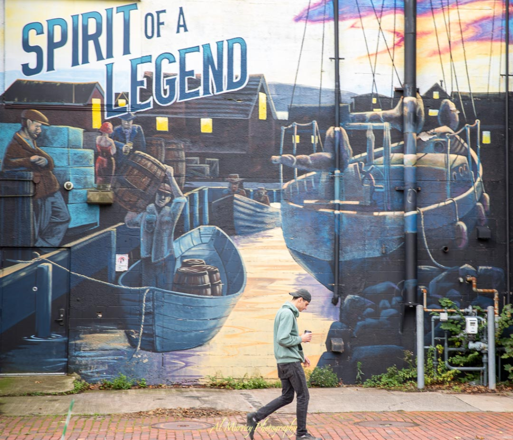 Colorful mural titled "Spirit of a Legend" depicting a historic maritime scene with dock workers, barrels, small boats, and a large sailing ship at sunset, painted on the side of a building. A man walks by in modern clothes, contrasting the vintage harbor imagery. This mural adds local character and charm to a Halifax or Cape Breton romantic getaway.
