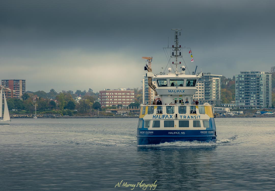 Halifax Transit ferry named "Vincent Coleman" sails across the harbor under moody grey skies, with modern waterfront buildings and a sailboat in the background. Couples and passengers stand on the open-air deck enjoying the scenic ride. A peaceful ferry ride like this adds charm to a Halifax romantic getaway.