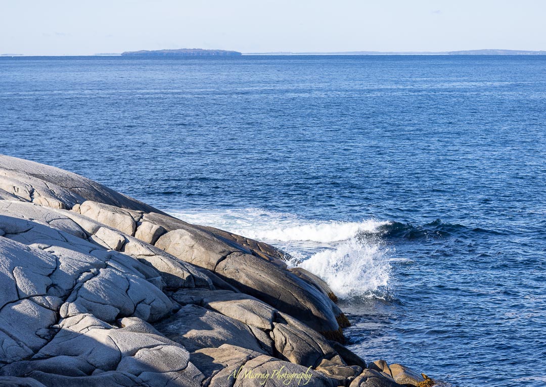 Waves crash against smooth, weathered granite rocks along the Atlantic shoreline under a clear blue sky, with distant islands visible on the horizon. The rugged coastal beauty offers a serene and dramatic setting for a Halifax or Cape Breton romantic getaway.