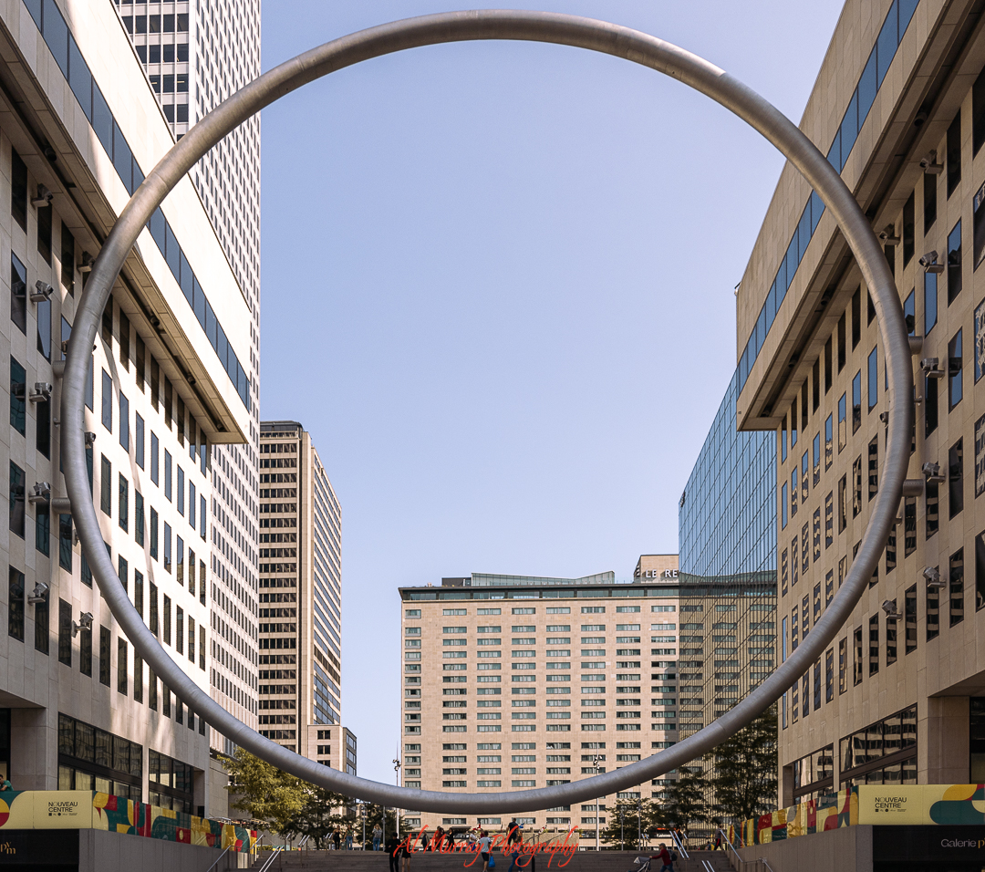 A massive metal ring sculpture frames a view of downtown Montreal’s modern architecture under a clear sky, creating a striking urban backdrop. This artistic landmark offers a unique photo spot and cultural stop on a Montreal Valentine's Day itinerary for couples.
