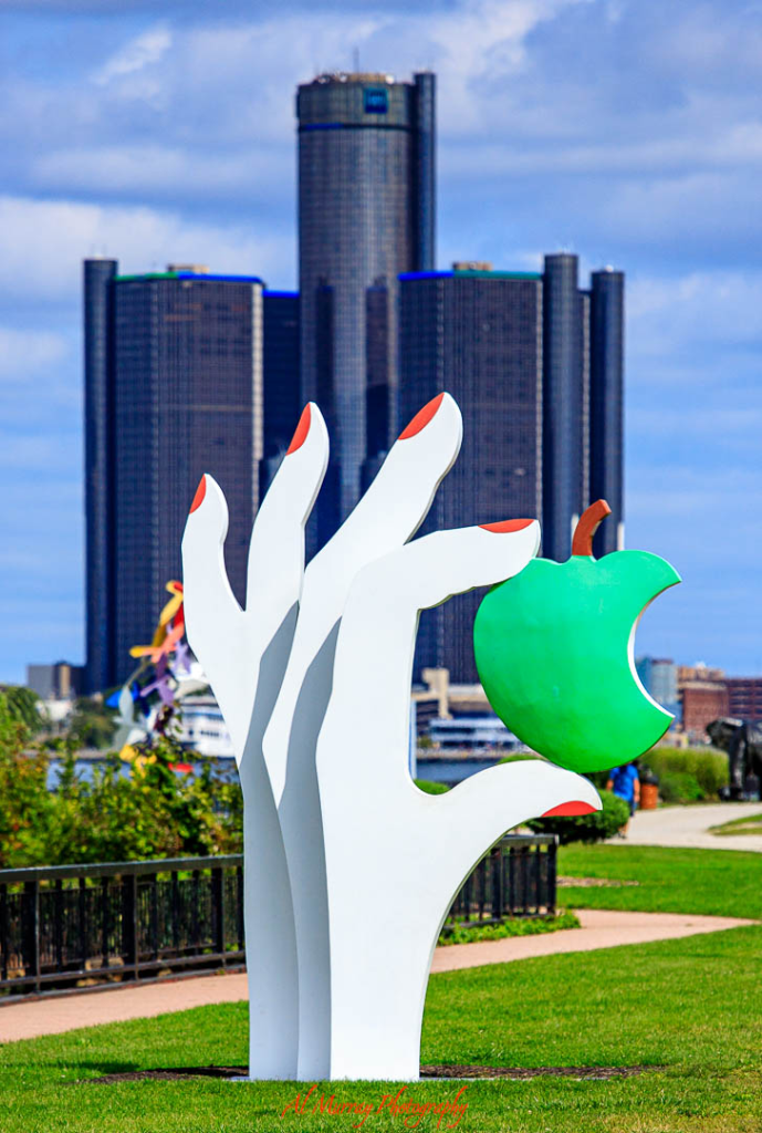 A playful sculpture of three white hands with red fingernails holding a bright green bitten apple stands along Windsor's waterfront park, with the Detroit skyline rising in the background. This vibrant public art stop offers a scenic and leisurely stroll perfect for older adults looking for relaxing things to do in Windsor Essex County in spring.
