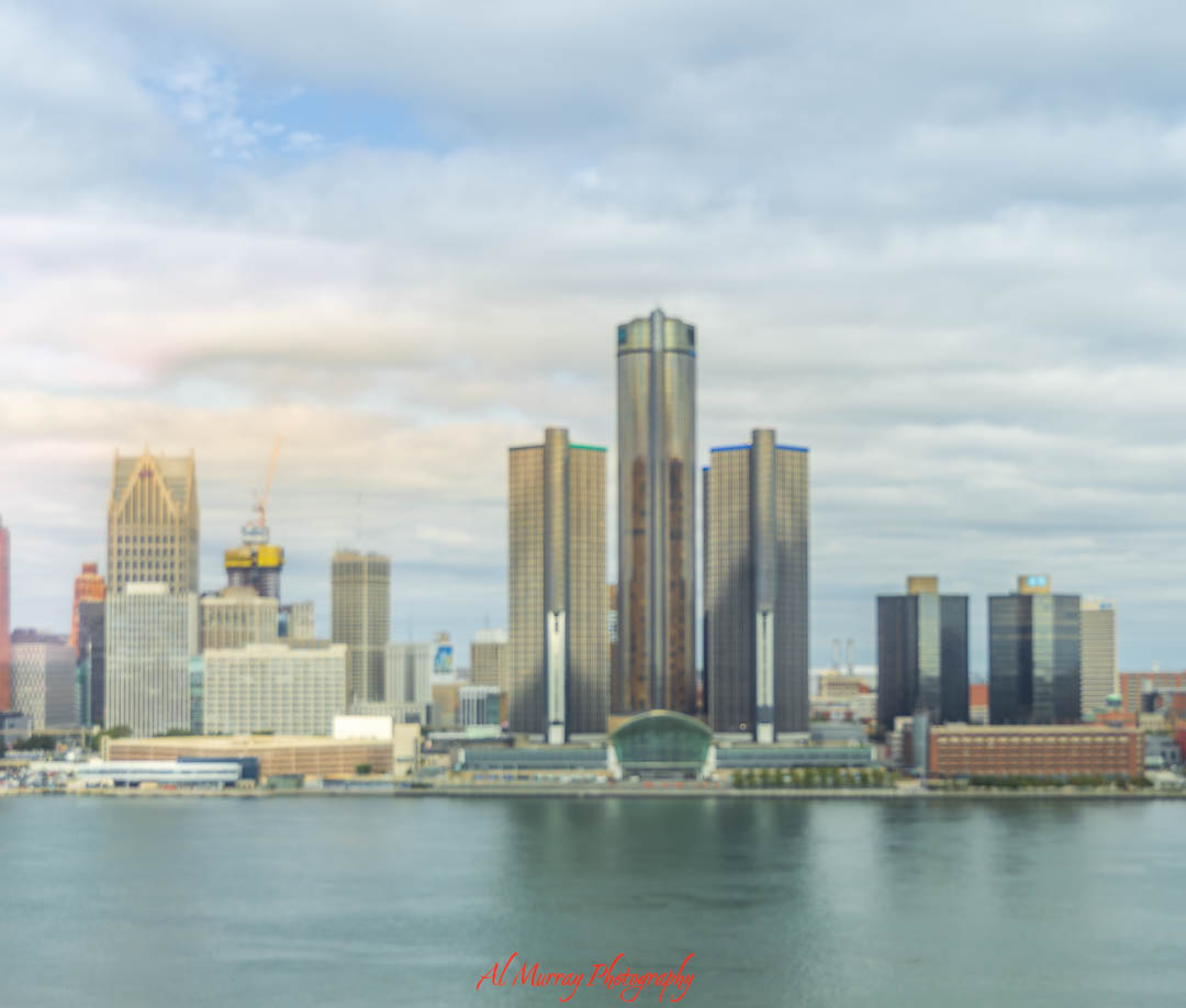 A soft-focus view of the Detroit skyline as seen across the Detroit River from Windsor Ontario, with glassy water and overcast skies creating a calm spring atmosphere. While not a vineyard scene itself, this urban vantage point marks the start of many spring wine tours in Essex County, where city meets countryside charm.
