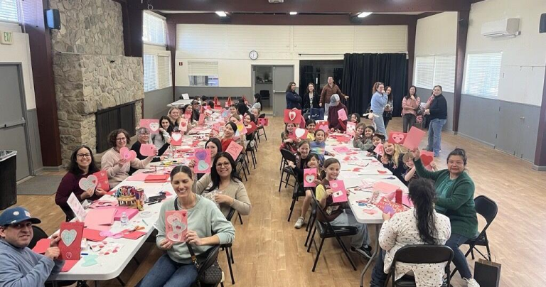 Group of adults and children sitting around a long table in a community center, proudly holding up handmade Valentine’s Day cards with pink and red hearts. The cheerful crafting event captures a warm, social atmosphere that contrasts with the experience of solo seniors on Valentine’s Day in Canada.