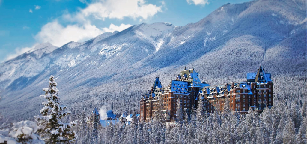 The Fairmont Banff Springs Hotel stands majestically among snow-covered pine trees with the Rocky Mountains rising in the background under a bright blue sky. This castle-like resort nestled in a winter wonderland is one of the most romantic places to visit in Canada in February.