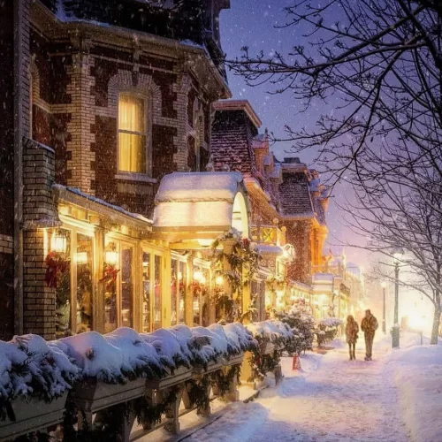 Adirondack chairs circle a glowing fire pit in the snow at dusk, overlooking a frozen lake and distant lights, creating a cozy winter scene perfect for a Niagara-on-the-Lake or Muskoka romantic getaway. The warm fire contrasts with the snowy setting, inviting couples to relax under the trees.