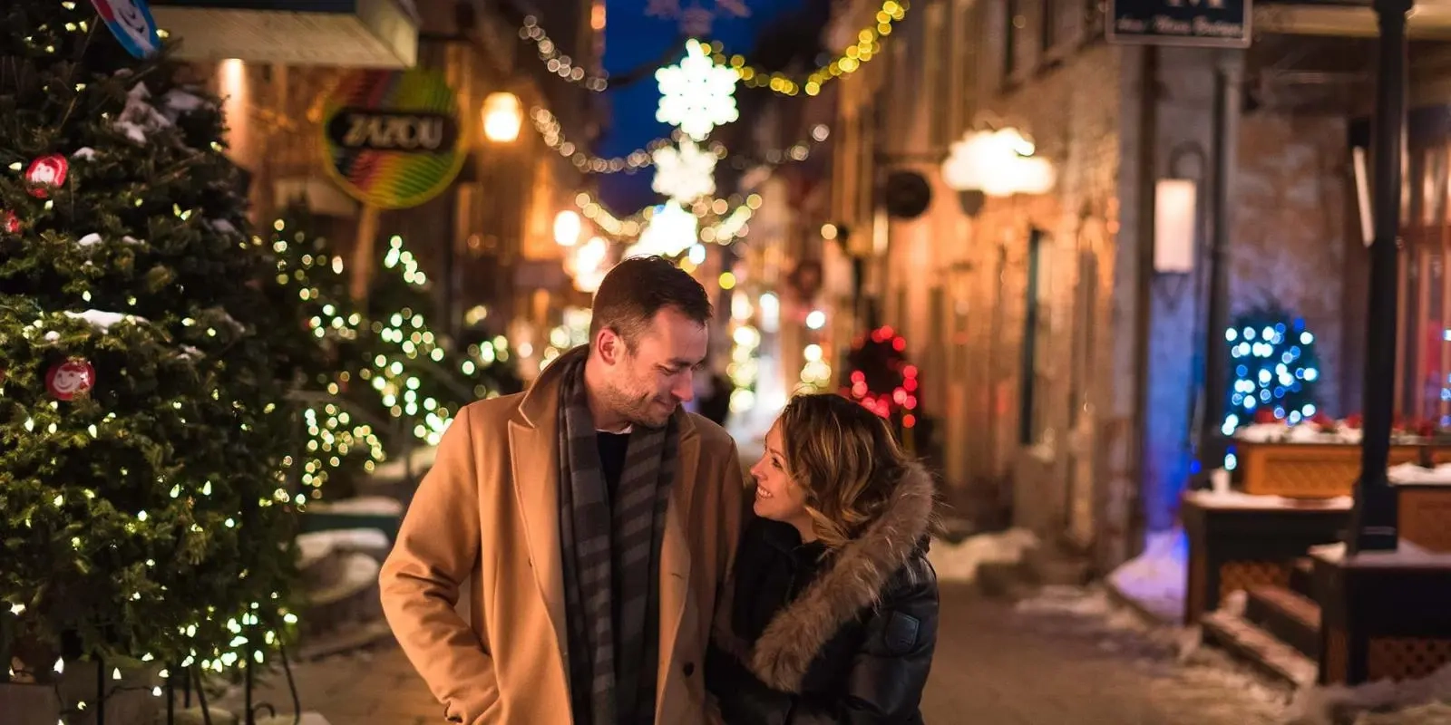 Couple smiles warmly at each other while strolling through a festive, snow-dusted street in Old Quebec, illuminated by holiday lights and decorated Christmas trees. The cozy, intimate atmosphere makes this one of the most romantic places in Canada.