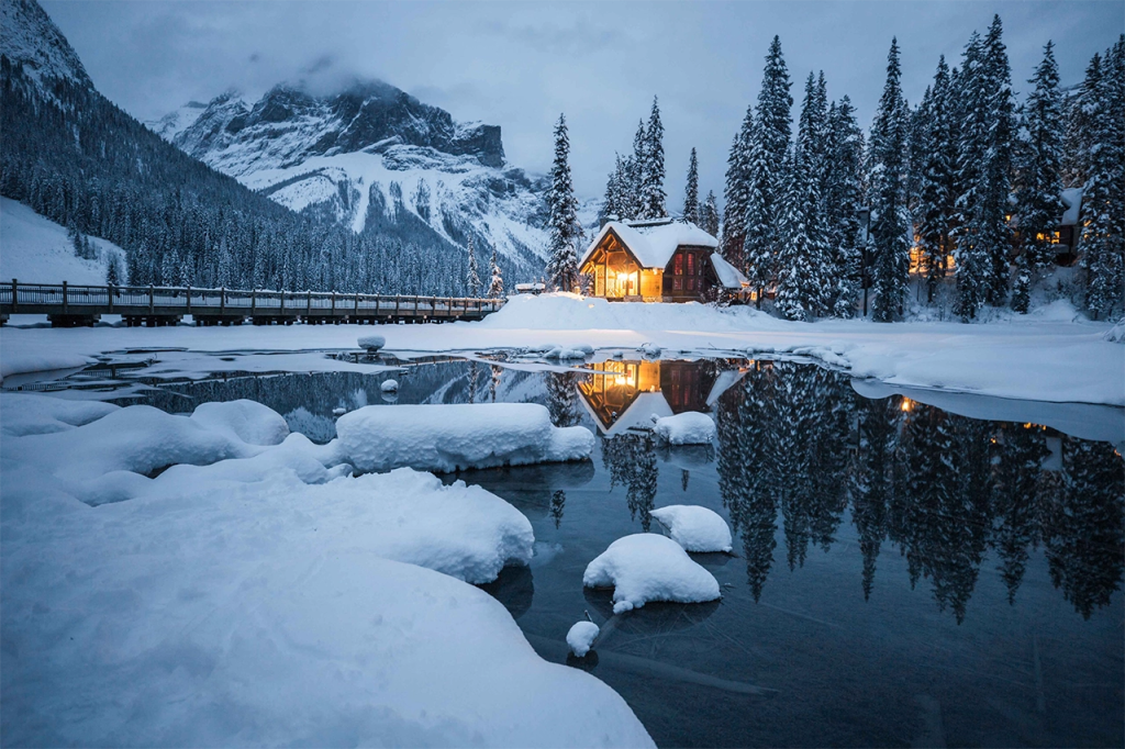 A glowing cabin sits nestled in a snowy forest beside a partially frozen lake, with warm light reflecting on the still water and snow-covered mountains in the background. This peaceful, secluded setting in the Rockies is one of the most romantic places in Canada for couples seeking winter magic.