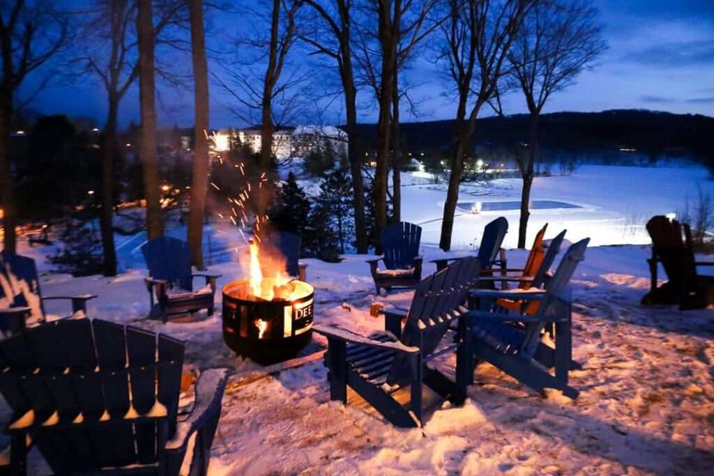 Adirondack chairs surround a crackling fire pit on a snowy hilltop at twilight, with a frozen lake and cozy resort lights glowing in the distance. This peaceful winter scene captures the charm of a Muskoka romantic getaway for couples seeking warmth and quiet under the stars.