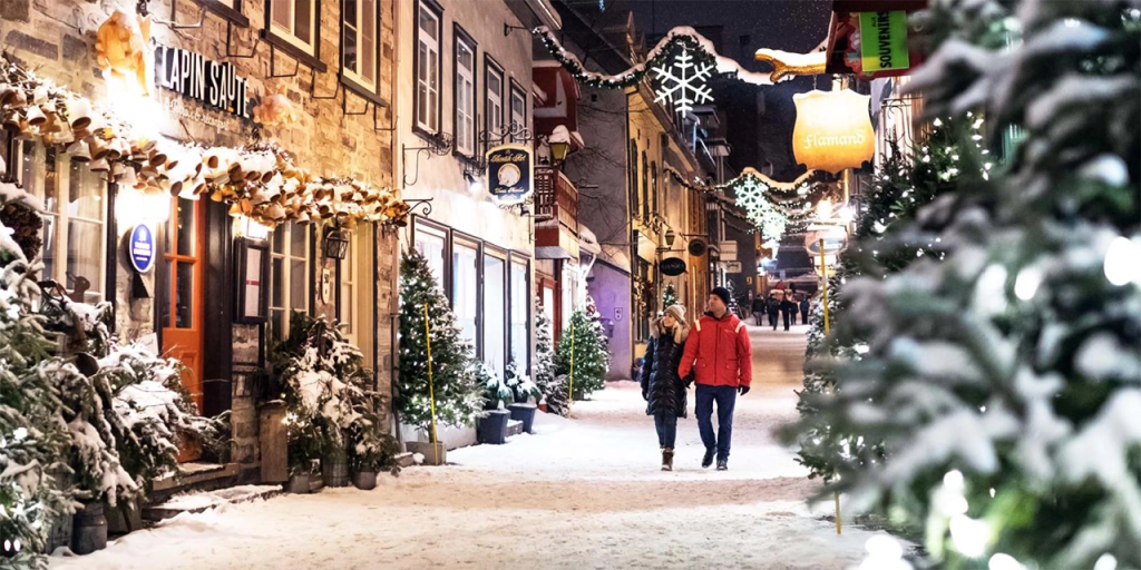 Couple walks hand-in-hand down a snow-covered, festively lit street in Old Quebec, surrounded by twinkling lights, decorated storefronts, and holiday garlands. The cozy winter charm and European ambiance make this one of the most romantic places in Canada for Valentine’s Day.
