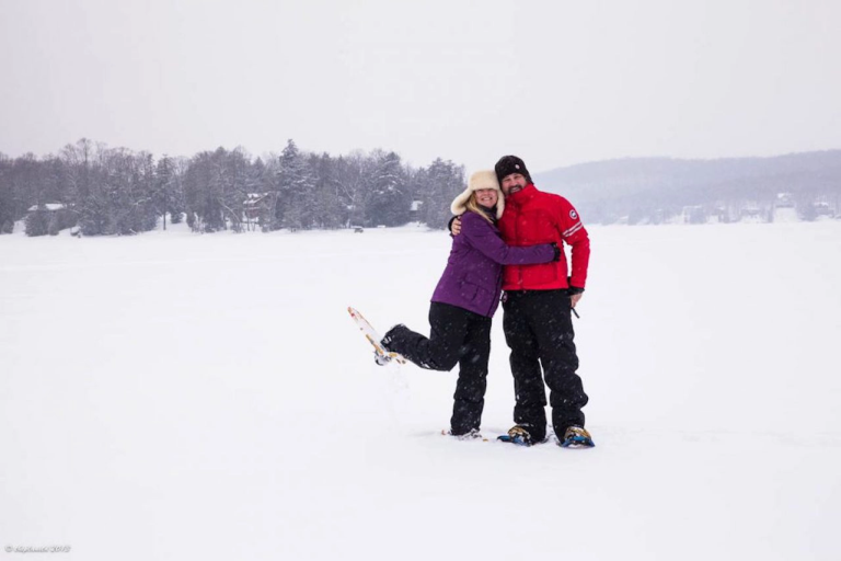 A smiling couple in winter gear embraces while snowshoeing on a frozen, snow-covered lake, surrounded by forested shoreline and softly falling snow. This joyful moment captures the playful spirit of a Niagara-on-the-Lake or Muskoka romantic getaway.
