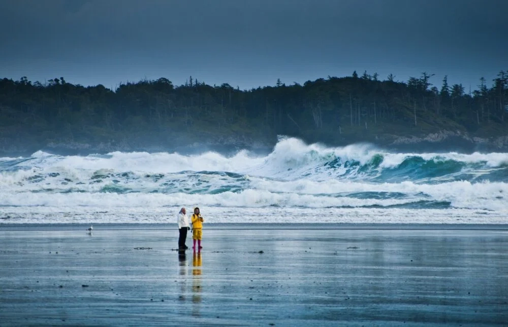 Two people stand together on a reflective, wet beach in front of dramatic crashing waves and a misty forested coastline under moody skies. The wild, rugged beauty of the scene captures the untamed romance of Tofino, one of the most romantic places in Canada.