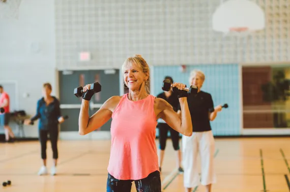 Smiling senior woman lifting dumbbells in a group fitness class inside a gym, surrounded by other older adults exercising with hand weights. Staying active through community workouts is one of many healthy solo seniors Valentine’s Day activities that boost both physical and emotional well-being.
