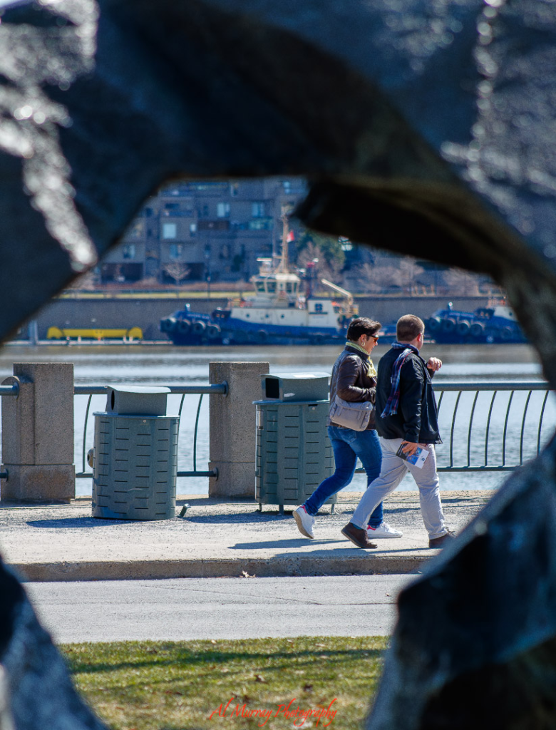 A man and woman walk along the waterfront in Montreal, framed through an abstract sculpture with a tugboat and calm river in the background. Peaceful strolls by the Old Port offer one of the quiet things to do in Montreal, ideal for relaxation and solo reflection.