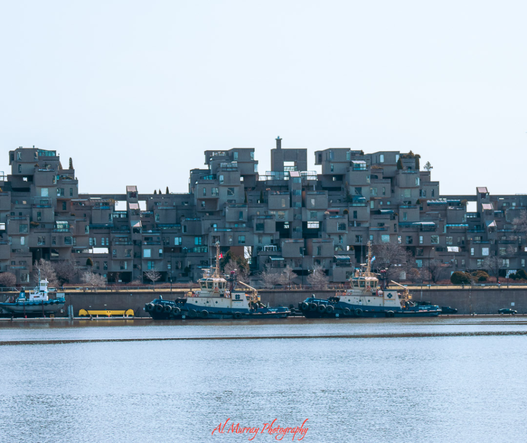 View of Habitat 67, a striking modular housing complex along the Montreal waterfront, with tugboats docked in the foreground. Admiring this architectural landmark from the riverside offers a peaceful, visually engaging outing—one of many low-impact things to do alone in Montreal for seniors.