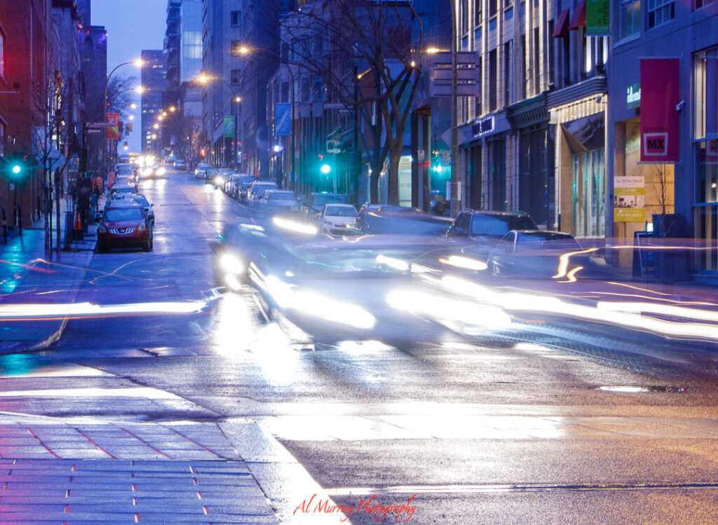 Long exposure shot of a downtown Montreal street at dusk, with blurred headlights from passing cars and reflections glistening on the wet pavement. Walking through the city’s vibrant core in the evening is one of many atmospheric things to do alone in Montreal.