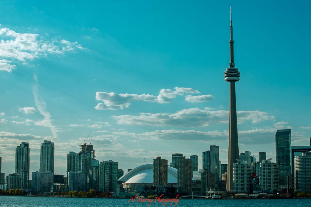 where to watch the world cup 2026 in Toronto Toronto skyline at sunset with the CN Tower rising above downtown skyscrapers and the waterfront in the foreground under a blue sky with scattered clouds. The cityscape highlights iconic gathering spots and potential Toronto fan zones World Cup 2026 visitors can explore while celebrating matches in the heart of the city.