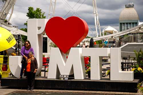 Two women pose in front of a large “I ❤️ MTL” sign at the Old Port in Montreal, with the Montreal Observation Wheel and dome of the Marché Bonsecours visible in the background. A popular photo spot for tourists, this area is one of many scenic places to explore solo in Montreal.