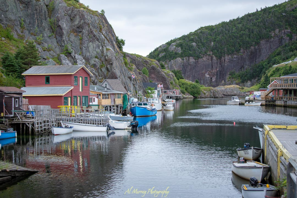 A peaceful harbor scene in Newfoundland with colorful fishing stages, small boats, and steep rocky cliffs under a cloudy spring sky. The quiet inlet and rustic charm make it a perfect stop on a Newfoundland spring road trip.