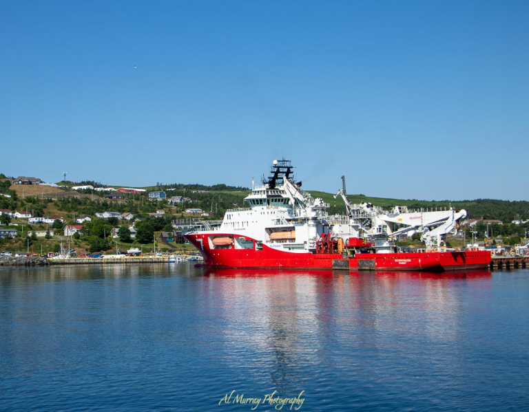 A bright red offshore vessel docked at a calm harbor with the green hills and colorful houses of Newfoundland in the background on a clear spring day. The image captures the vibrant waterfront and coastal charm typical of spring in St. John’s and Newfoundland.