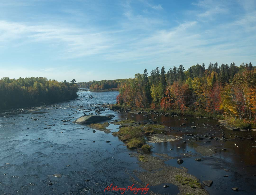 Wide landscape photo of a calm river winding through a forest filled with vibrant autumn foliage in shades of red, orange, and gold under a soft blue sky. The peaceful water and colorful trees capture the natural beauty travelers can experience on a Bay of Fundy road trip, highlighting scenic inland stops beyond the dramatic coastal tides.