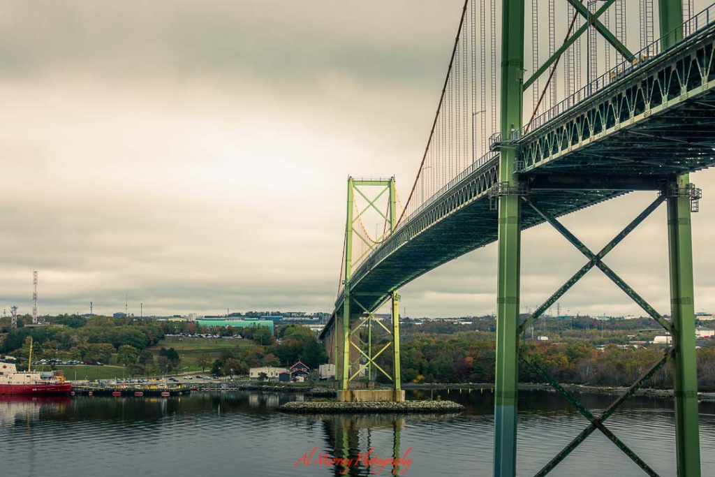 Wide angle photo of a large green suspension bridge stretching over calm harbor water with tree covered shoreline in the background under an overcast sky. The towering steel structure and reflective water capture a scenic departure point for a Peggy’s Cove day trip from Halifax, highlighting the coastal views travelers experience along the way.
