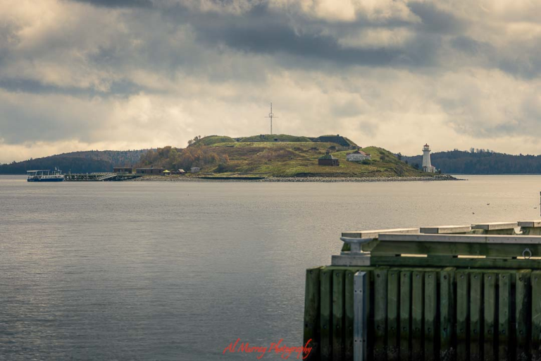 Wide landscape photo of a small island with a white lighthouse, grassy hill, and scattered buildings surrounded by calm harbor waters under a cloudy sky. Viewed from a wooden pier in the foreground, the peaceful coastal scene reflects a scenic stop on a Maritimes driving tour for seniors from Halifax, showcasing accessible waterfront views and classic Atlantic charm.